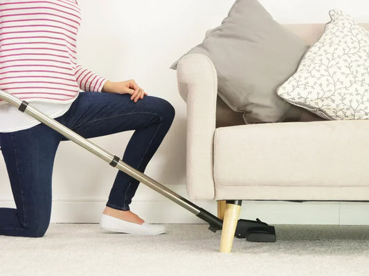 A person kneeling on a carpeted floor while using a vacuum cleaner wand to reach and clean underneath a light beige sofa.